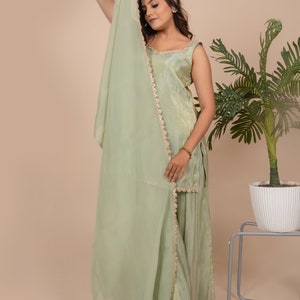 May include: A light sage green sleeveless tunic and matching wide-leg trousers set, paired with a sheer, lightweight dupatta scarf. The tunic and scarf feature delicate gold-toned metallic trim along the edges, creating a cohesive ensemble.