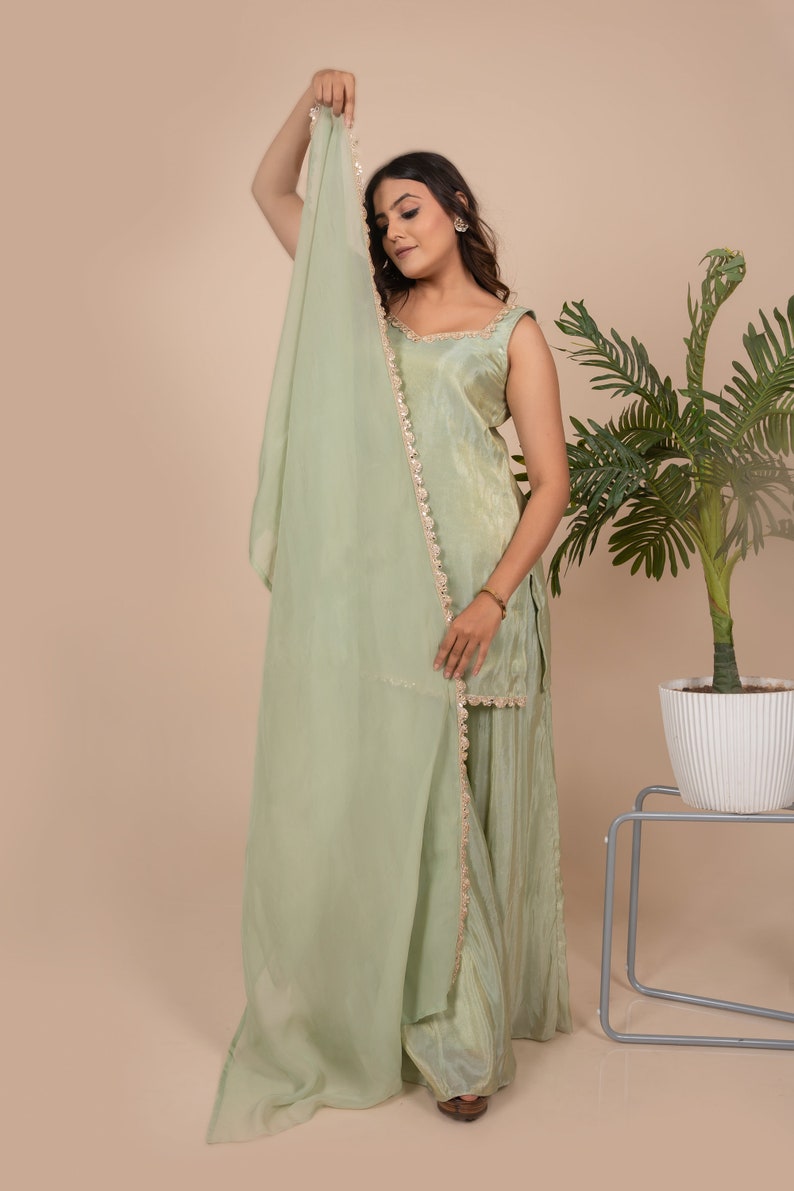 May include: A light sage green sleeveless tunic and matching wide-leg trousers set, paired with a sheer, lightweight dupatta scarf. The tunic and scarf feature delicate gold-toned metallic trim along the edges, creating a cohesive ensemble.