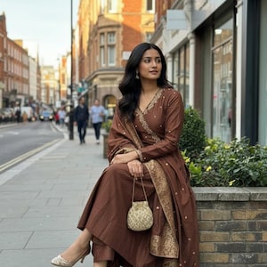 May include: A brown salwar kameez set with gold embroidery. The outfit includes a long-sleeved top, pants, and a matching scarf. A small, gold-colored purse is held in the hand. The person is wearing gold-colored heels.