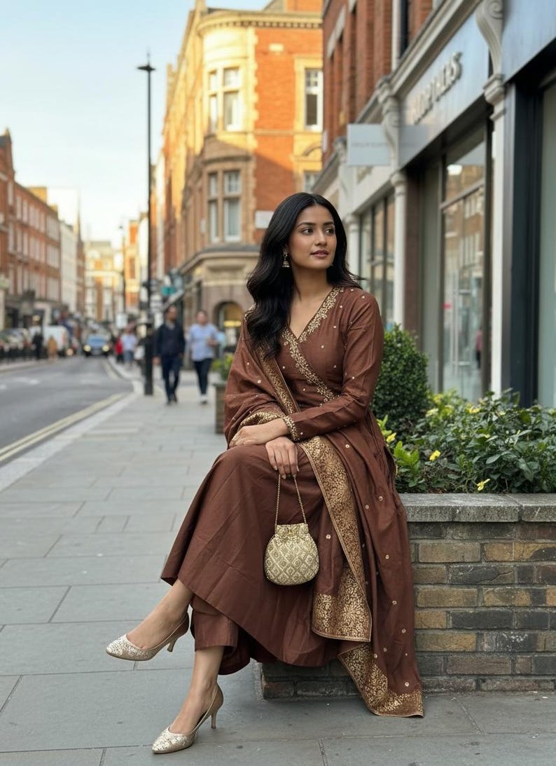 May include: A brown salwar kameez set with gold embroidery. The outfit includes a long-sleeved top, pants, and a matching scarf. A small, gold-colored purse is held in the hand. The person is wearing gold-colored heels.