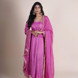 May include: A pink long-sleeved dress with gold trim and a matching pink dupatta. The dress has a square neckline and a fitted bodice. The dupatta is draped over the shoulders and has a fringed border.