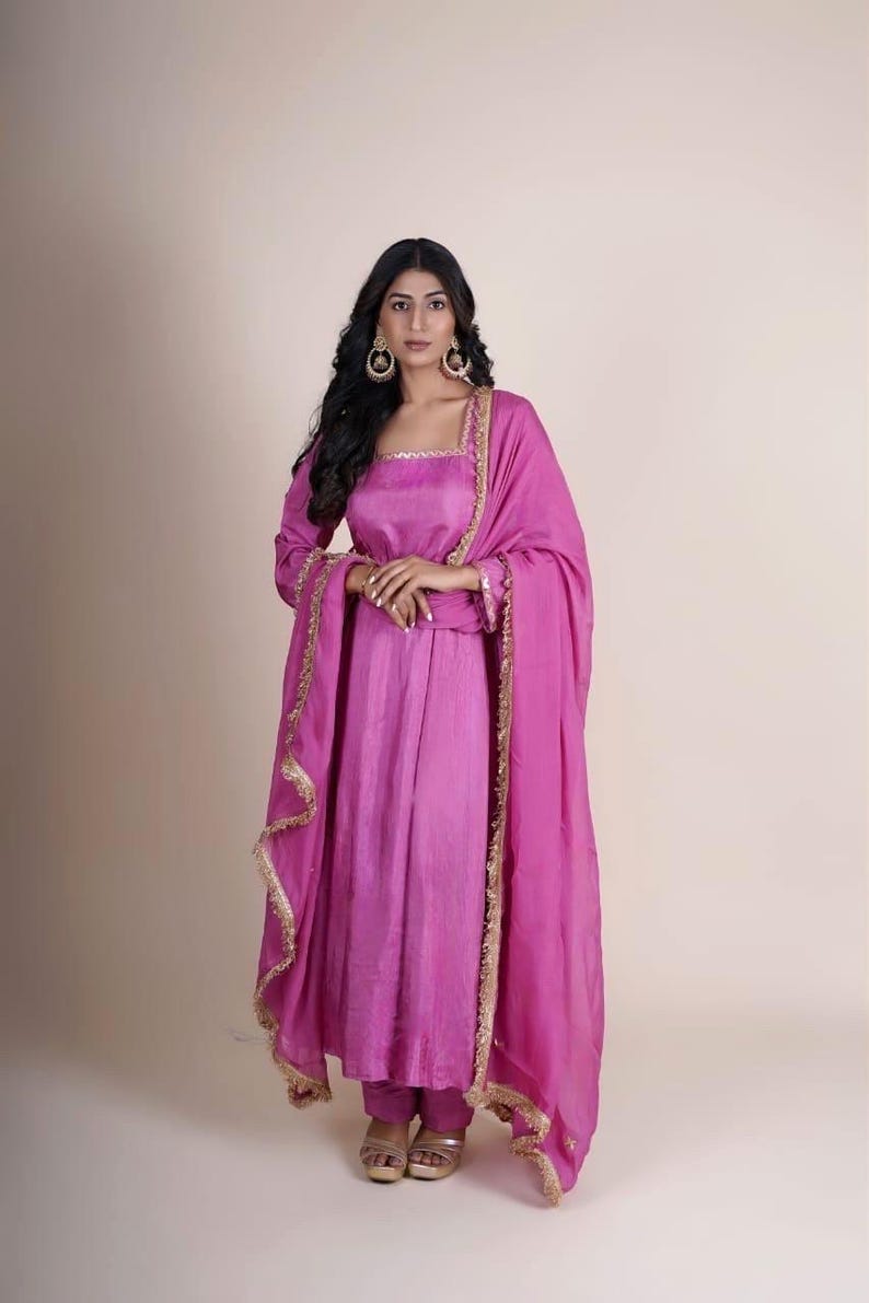 May include: A pink long-sleeved dress with gold trim and a matching pink dupatta. The dress has a square neckline and a fitted bodice. The dupatta is draped over the shoulders and has a fringed border.
