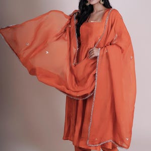 May include: An orange salwar kameez set. The ensemble includes a long tunic, matching pants, and a sheer dupatta. The dupatta and tunic are trimmed with silver detailing. The outfit is made of a flowing fabric.