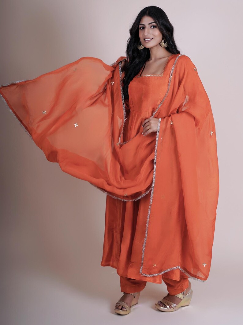 May include: An orange salwar kameez set. The ensemble includes a long tunic, matching pants, and a sheer dupatta. The dupatta and tunic are trimmed with silver detailing. The outfit is made of a flowing fabric.