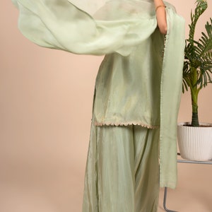 May include: A light sage green ensemble consisting of a sleeveless tunic with a scalloped hem and wide-leg trousers. The outfit is paired with a matching sheer, lightweight dupatta draped across the shoulders, featuring a subtle sheen and delicate texture.