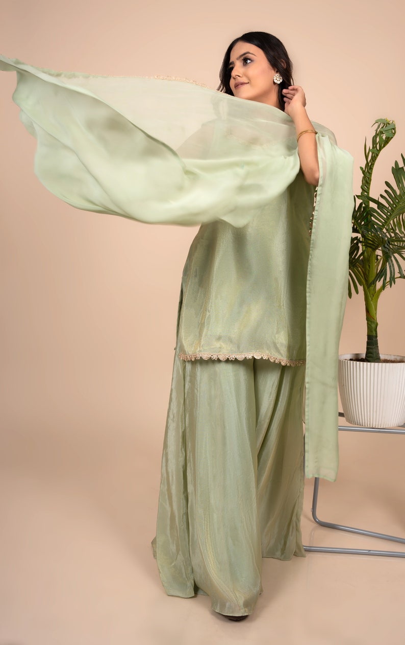 May include: A light sage green ensemble consisting of a sleeveless tunic with a scalloped hem and wide-leg trousers. The outfit is paired with a matching sheer, lightweight dupatta draped across the shoulders, featuring a subtle sheen and delicate texture.