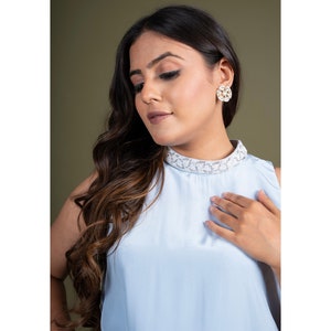 May include: A woman wearing a light blue sleeveless top with a white lace collar and a pair of gold and white floral earrings.