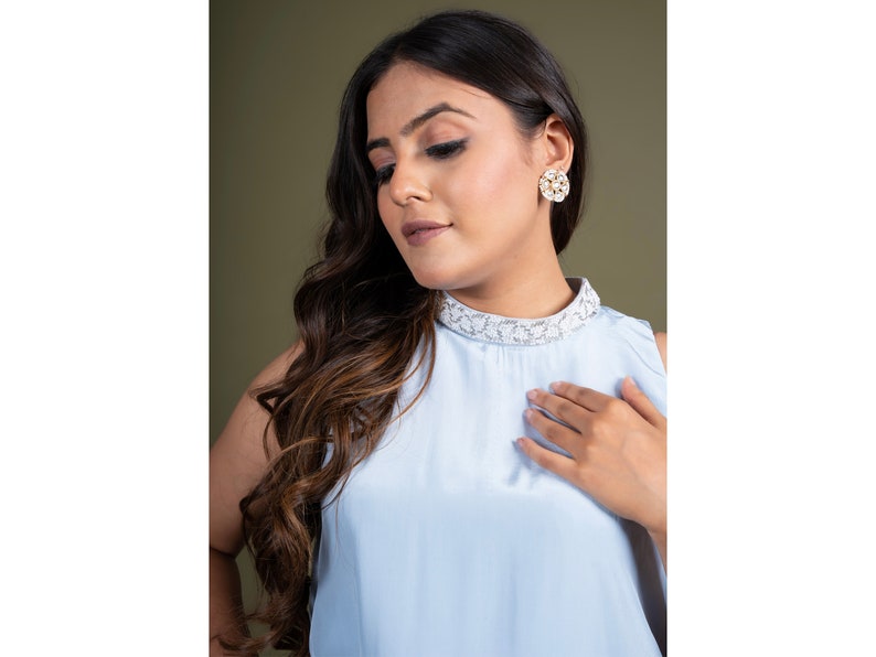 May include: A woman wearing a light blue sleeveless top with a white lace collar and a pair of gold and white floral earrings.