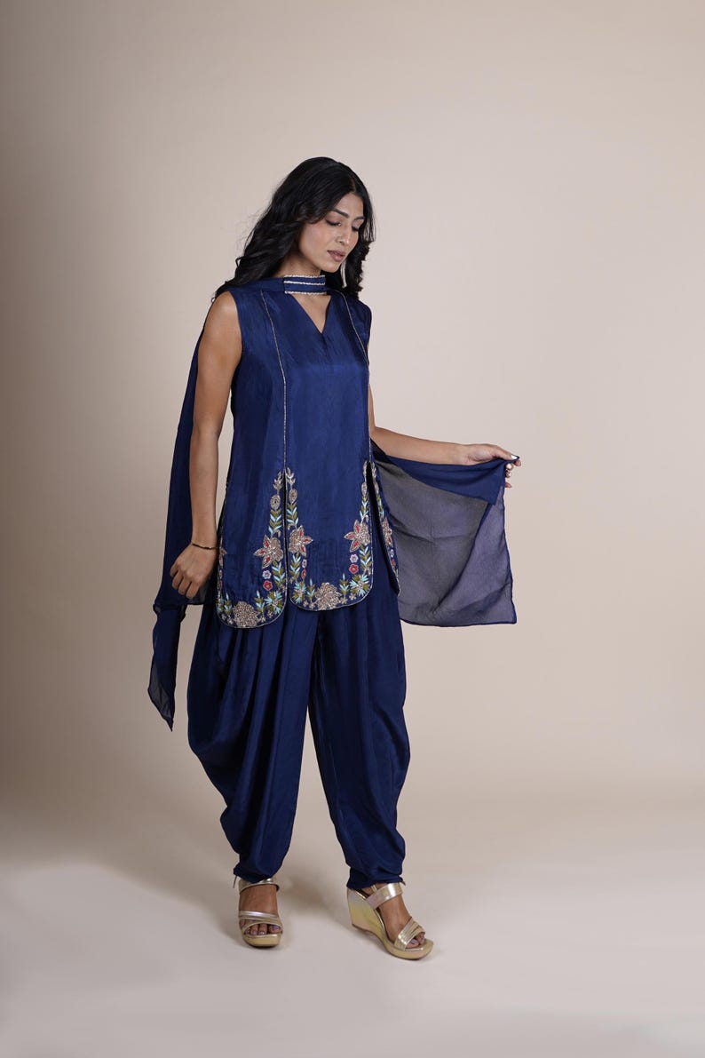 May include: A woman wearing a navy blue salwar kameez with floral embroidery. The outfit includes a long, sleeveless top with a V-neckline and a pair of matching pants.
