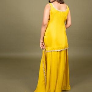 May include: A bright yellow sleeveless tunic paired with a matching floor-length skirt. The tunic features a scoop neckline and a hemline adorned with intricate white scalloped lace trim. A long, flowing yellow fabric drape extends from the side.
