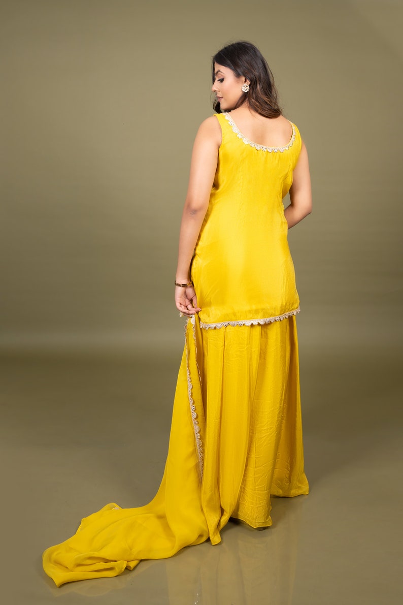 May include: A bright yellow sleeveless tunic paired with a matching floor-length skirt. The tunic features a scoop neckline and a hemline adorned with intricate white scalloped lace trim. A long, flowing yellow fabric drape extends from the side.