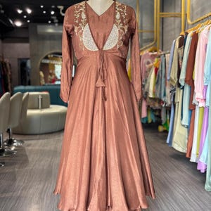 May include: A copper-colored, long-sleeved dress and matching pants set on a mannequin. The dress features a V-neck with embroidered detailing and a tie closure. The outfit is displayed in a boutique setting, with other garments visible in the background.