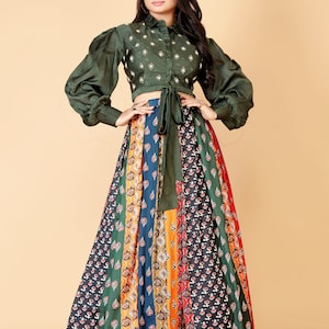 May include: An olive green crop top with long, puffed sleeves and a tie at the waist, paired with a long, patterned skirt. The skirt features vertical panels of various colors and floral designs. The outfit is suitable for special occasions.