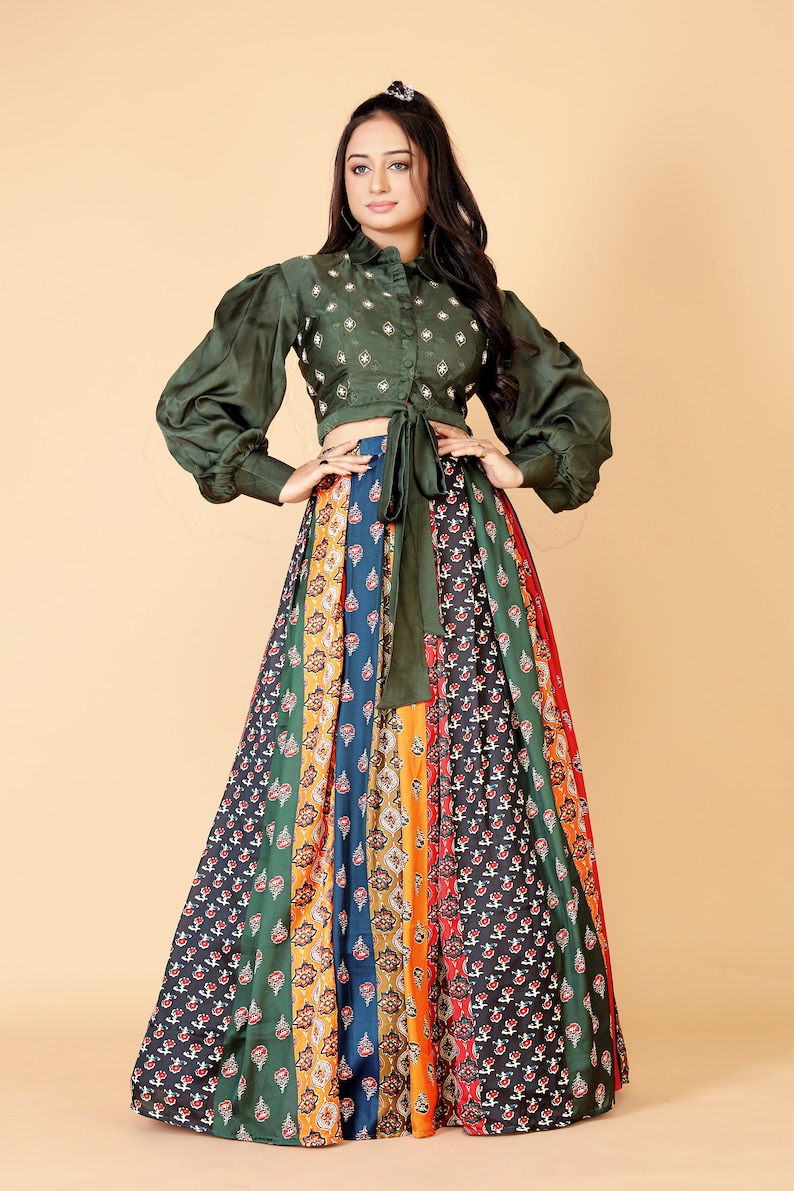 May include: An olive green crop top with long, puffed sleeves and a tie at the waist, paired with a long, patterned skirt. The skirt features vertical panels of various colors and floral designs. The outfit is suitable for special occasions.