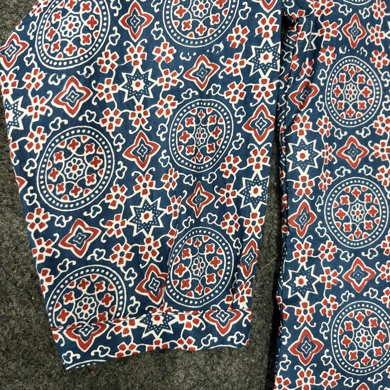 Op de afbeelding: Blauwe stof met een rood en wit geometrisch patroon. Het patroon bevat cirkels, sterren en vierkanten.