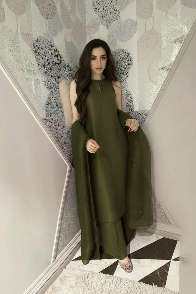 May include: Olive green sleeveless dress and matching pants with a flowing shawl. The outfit has a keyhole neckline and is made of a smooth, solid fabric. The ensemble is worn with embellished shoes.