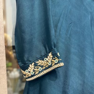 Pu&ograve; includere: Abito color verde acqua con maniche lunghe. Il polsino della manica presenta ricami dorati con dettagli bianchi e verdi. L'abito ha una silhouette fluida e sembra essere realizzato in un materiale setoso. Il corpetto &egrave; delicatamente ricamato.