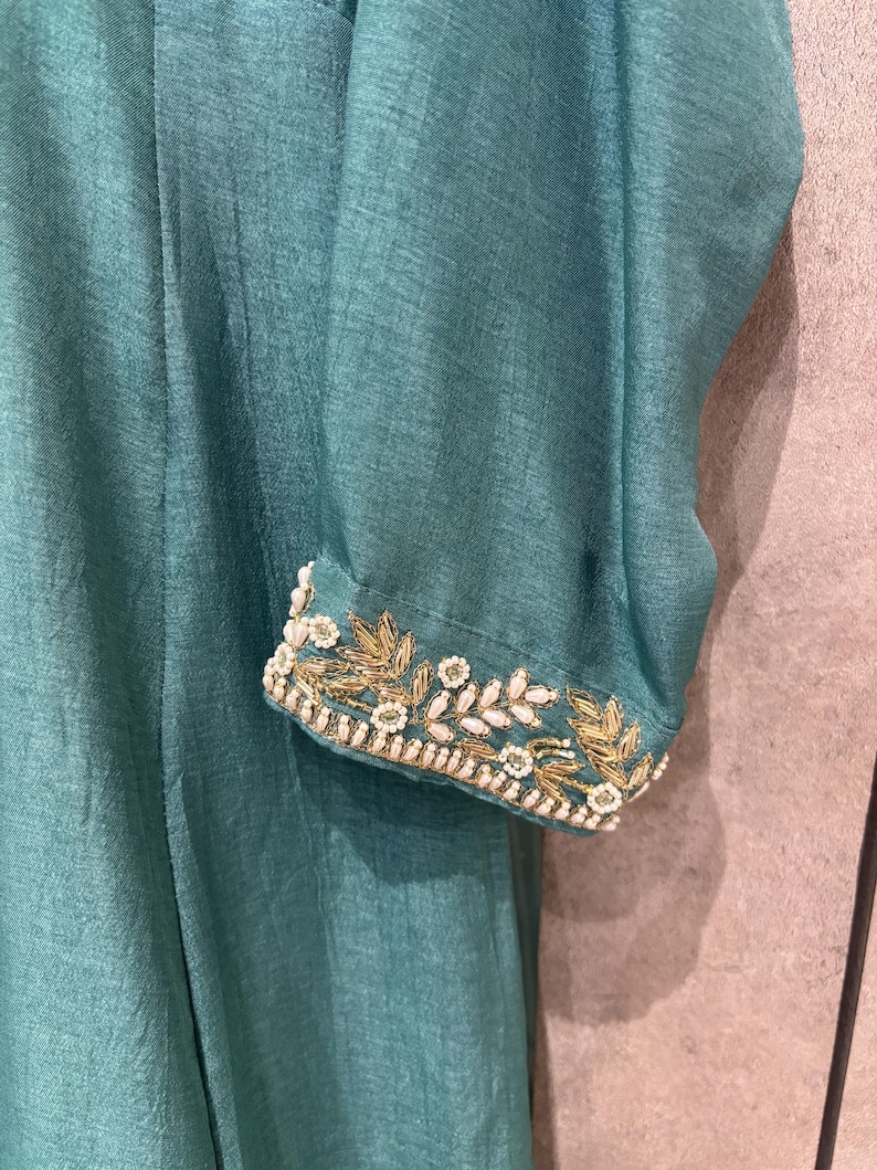 Pu&ograve; includere: Primo piano di un capo color verde acqua con manica lunga. Il polsino della manica &egrave; adornato da ricami dorati e bianchi intricati. Il tessuto ha una lucentezza sottile, che suggerisce un materiale lussuoso.