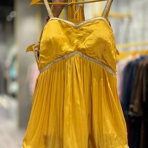 Puede incluir: Una blusa amarilla mostaza sin mangas con escote de coraz&oacute;n y falda con vuelo. La blusa tiene un ribete plateado alrededor del escote y la falda tiene un encaje plateado.