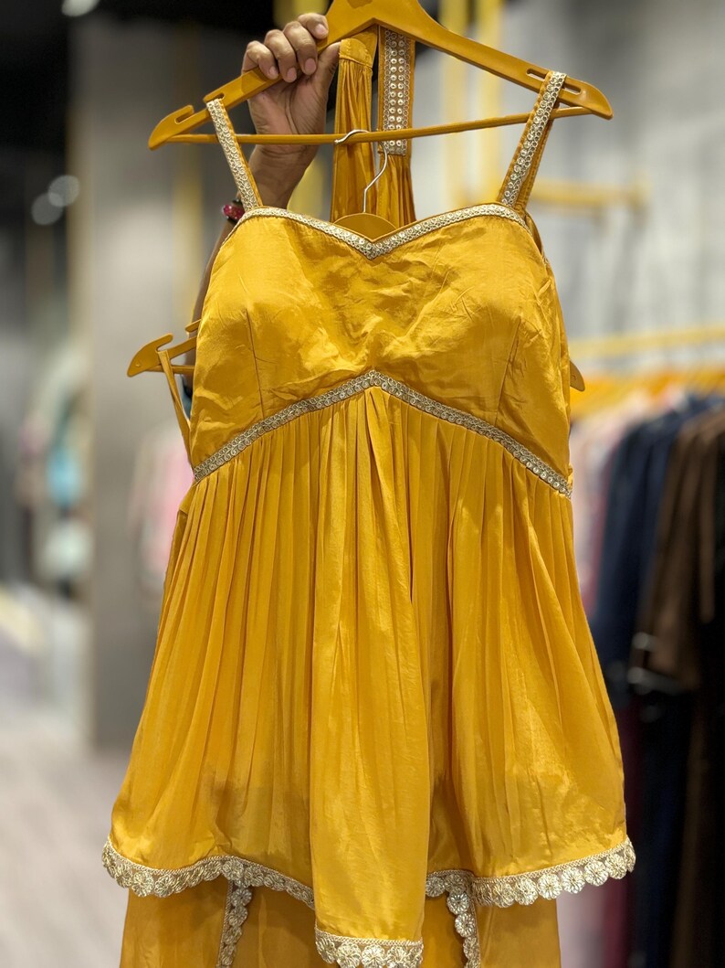 Puede incluir: Una blusa amarilla mostaza sin mangas con escote de coraz&oacute;n y falda con vuelo. La blusa tiene un ribete plateado alrededor del escote y la falda tiene un encaje plateado.