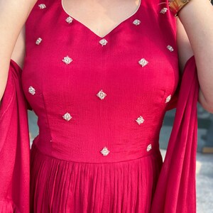 Pu&ograve; includere: Un abito maxi color granata con scollo a V e impreziosito da piccole paillettes bianche a forma di diamante. L'abito ha una gonna fluida e maniche a campana.