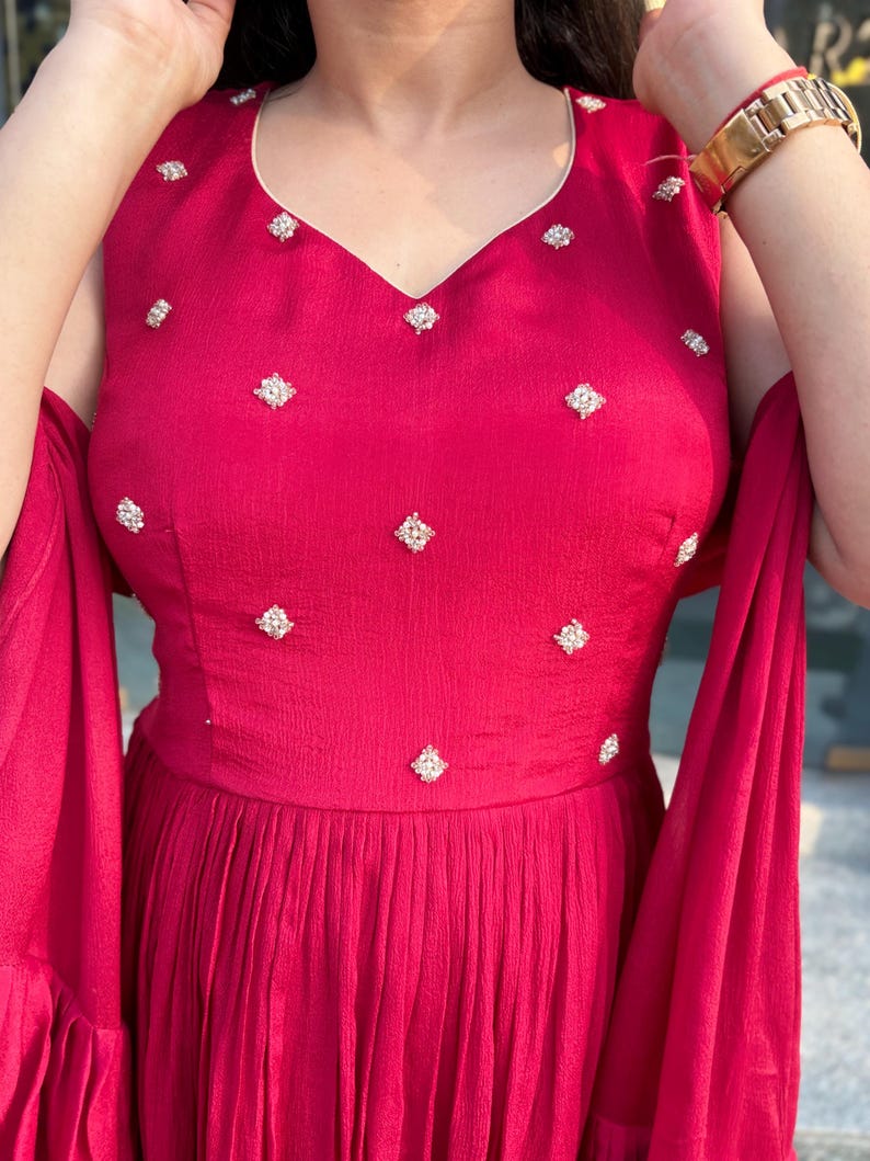 Pu&ograve; includere: Un abito maxi color granata con scollo a V e impreziosito da piccole paillettes bianche a forma di diamante. L'abito ha una gonna fluida e maniche a campana.