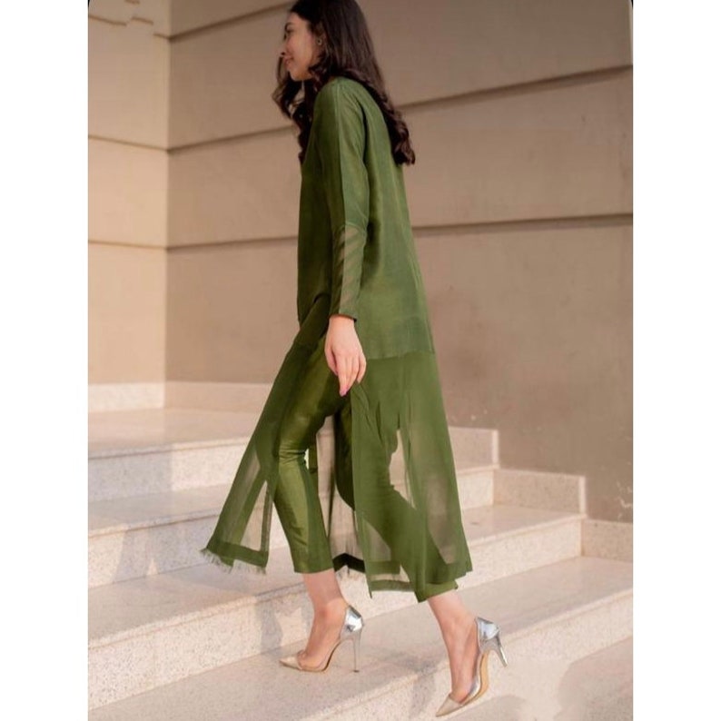 K&ouml;nnte beinhalten: Eine Frau tr&auml;gt eine lange, olivgr&uuml;ne Tunika mit einer passenden Hose. Die Tunika hat einen Schlitz vorne und die Hose ist kurz. Sie tr&auml;gt silberne High Heels.