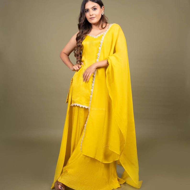 May include: A bright yellow sleeveless kurta paired with matching wide-leg sharara pants and a long, flowing dupatta. The ensemble features delicate white lace trim along the neckline, the side of the kurta, and the edges of the dupatta.