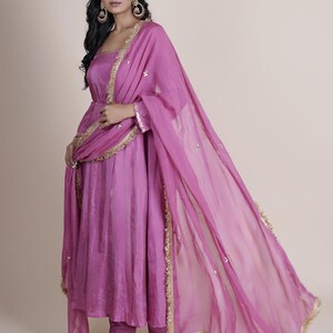 May include: A magenta-colored Indian salwar kameez suit. The suit features a long tunic top, matching pants, and a sheer dupatta scarf. Gold trim accents the edges of the dupatta and the neckline of the tunic. The outfit is made of a flowing fabric.