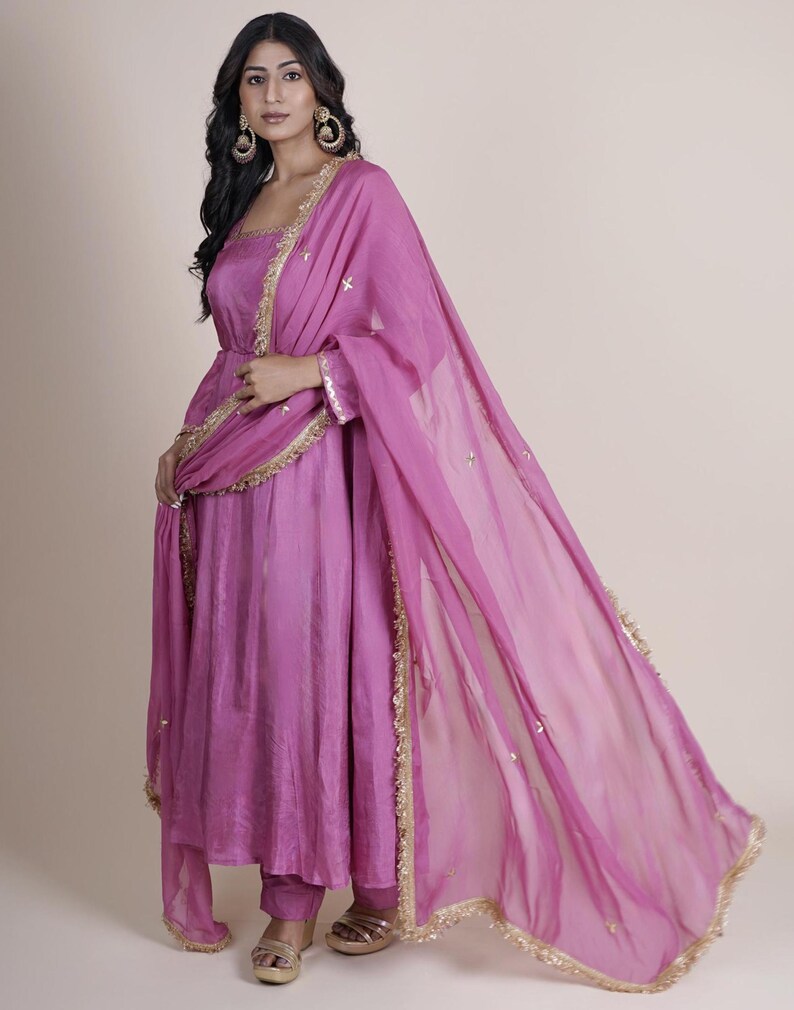 May include: A magenta-colored Indian salwar kameez suit. The suit features a long tunic top, matching pants, and a sheer dupatta scarf. Gold trim accents the edges of the dupatta and the neckline of the tunic. The outfit is made of a flowing fabric.