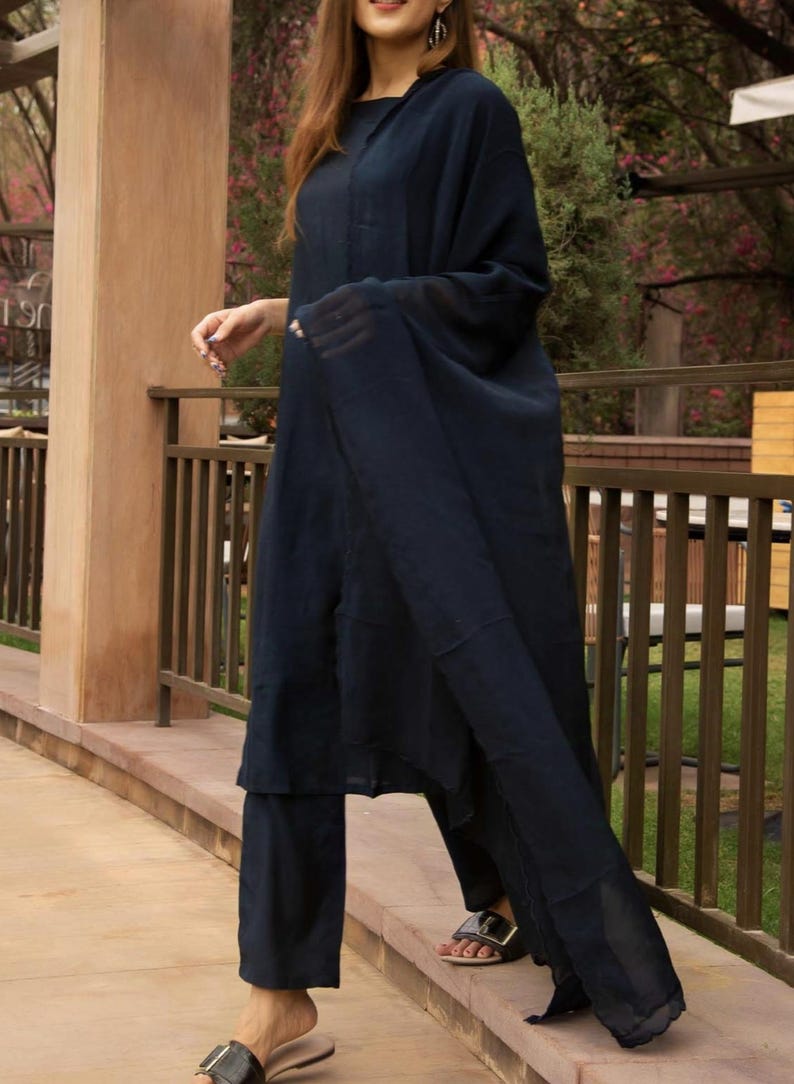 May include: A woman wearing a navy blue three-piece outfit, including a long-sleeved top, pants, and a sheer dupatta. The outfit is made of a soft, flowing fabric. The woman is standing on a stone path.