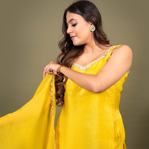 May include: A bright yellow sleeveless tunic with a matching sheer dupatta, both featuring delicate scalloped gold embroidery along the edges. The outfit is accessorized with round gold stud earrings and thin gold bangles on the wrist.