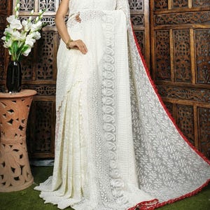 May include: A white saree with intricate floral embroidery and a red border. The saree is draped over a person, showcasing the delicate lace-like patterns. The background features a wooden screen and a vase of white flowers.