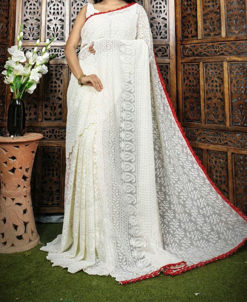 May include: A white saree with intricate floral embroidery and a red border. The saree is draped over a person, showcasing the delicate lace-like patterns. The background features a wooden screen and a vase of white flowers.