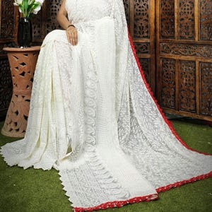 May include: A white saree with intricate floral embroidery. The saree features a red border and trim. The fabric appears to be lightweight and flowing. The saree is draped over a person sitting on a green surface.