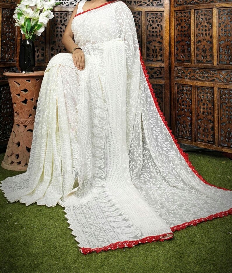 May include: A white saree with intricate floral embroidery. The saree features a red border and trim. The fabric appears to be lightweight and flowing. The saree is draped over a person sitting on a green surface.