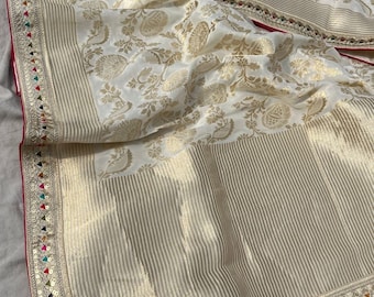 May include: A white silk saree with a gold floral pattern and a border of gold and colorful sequins. The saree has a pink and gold trim.