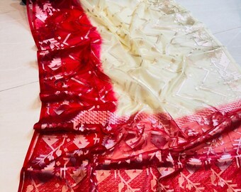 Puede incluir: Una tela transparente y fluida en un diseño de sari. El sari presenta un degradado de rojo a crema, con intrincados patrones y adornos. La parte roja tiene diseños florales y geométricos detallados.