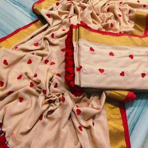 May include: A beige fabric with red heart embroidery, gold trim, and red tassels. The fabric is draped and folded, showcasing the design. The edges are trimmed with gold and red accents, creating a visually appealing textile.