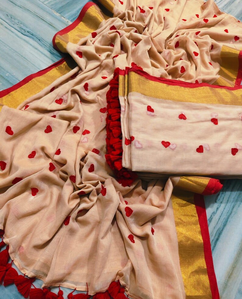 May include: A beige fabric with red heart embroidery, gold trim, and red tassels. The fabric is draped and folded, showcasing the design. The edges are trimmed with gold and red accents, creating a visually appealing textile.