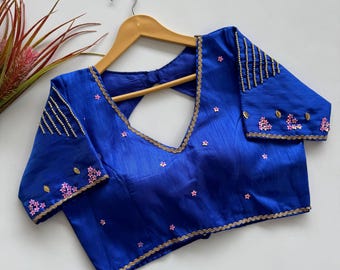 Op de afbeelding: Een koningsblauwe blouse met een V-hals en korte mouwen. De blouse is versierd met gouden randen en roze bloemenborduurwerk. De mouwen en halslijn zijn gedetailleerd met gouden kralen. Het hangt aan een houten hanger.