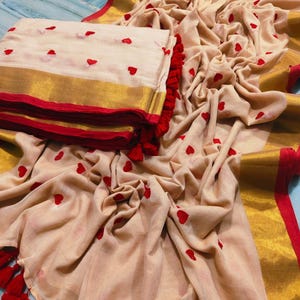 May include: A beige saree with red heart embroidery and a gold border. The saree is folded and stacked next to a draped portion, also featuring red heart accents and red tassels.