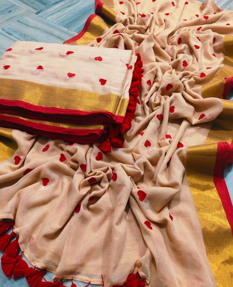 May include: A beige saree with red heart embroidery and a gold border. The saree is folded and stacked next to a draped portion, also featuring red heart accents and red tassels.