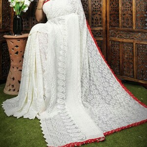 May include: A white saree with intricate floral embroidery and a red border. The saree is draped over a person, showcasing the delicate lace-like design. The background includes a wooden screen and a green surface.