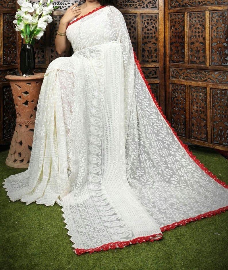 May include: A white saree with intricate floral embroidery and a red border. The saree is draped over a person, showcasing the delicate lace-like design. The background includes a wooden screen and a green surface.