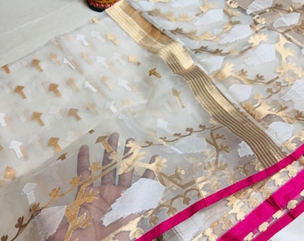 Puede incluir: Una tela blanca transparente con estampados florales dorados y blancos y un borde dorado. La tela tiene un borde rosa brillante. Una pequeña estatua de una figura sentada está en el fondo.