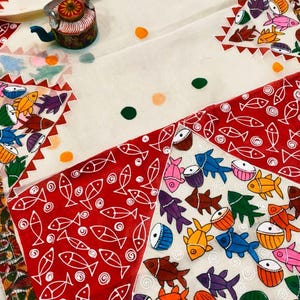 May include: A white textile with a colourful fish pattern and a decorative teapot. The fabric features red, white, and green accents, with a repeating fish motif in various colours. The teapot is small and ornate.