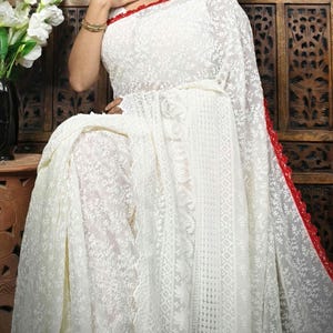 May include: White embroidered saree with a red border. The saree features intricate floral and geometric patterns throughout the fabric. The garment is draped over a person, showcasing the delicate design and texture.