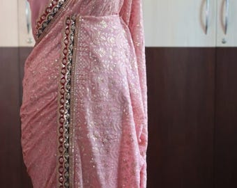 以下が含まれることがあります： 淡いピンク色のサリーで、同系色の繊細な花柄と装飾的な縁取りが特徴です。縁取りにはダークブルー、レッド、ゴールドのメタリックなアクセントが施されており、複雑な刺繍と小さな反射する円形の装飾があしらわれています。