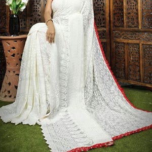 May include: A white saree with intricate embroidery and a red border. The saree is draped over a person, showcasing the delicate floral patterns and detailed design. The saree is made of a lightweight fabric.